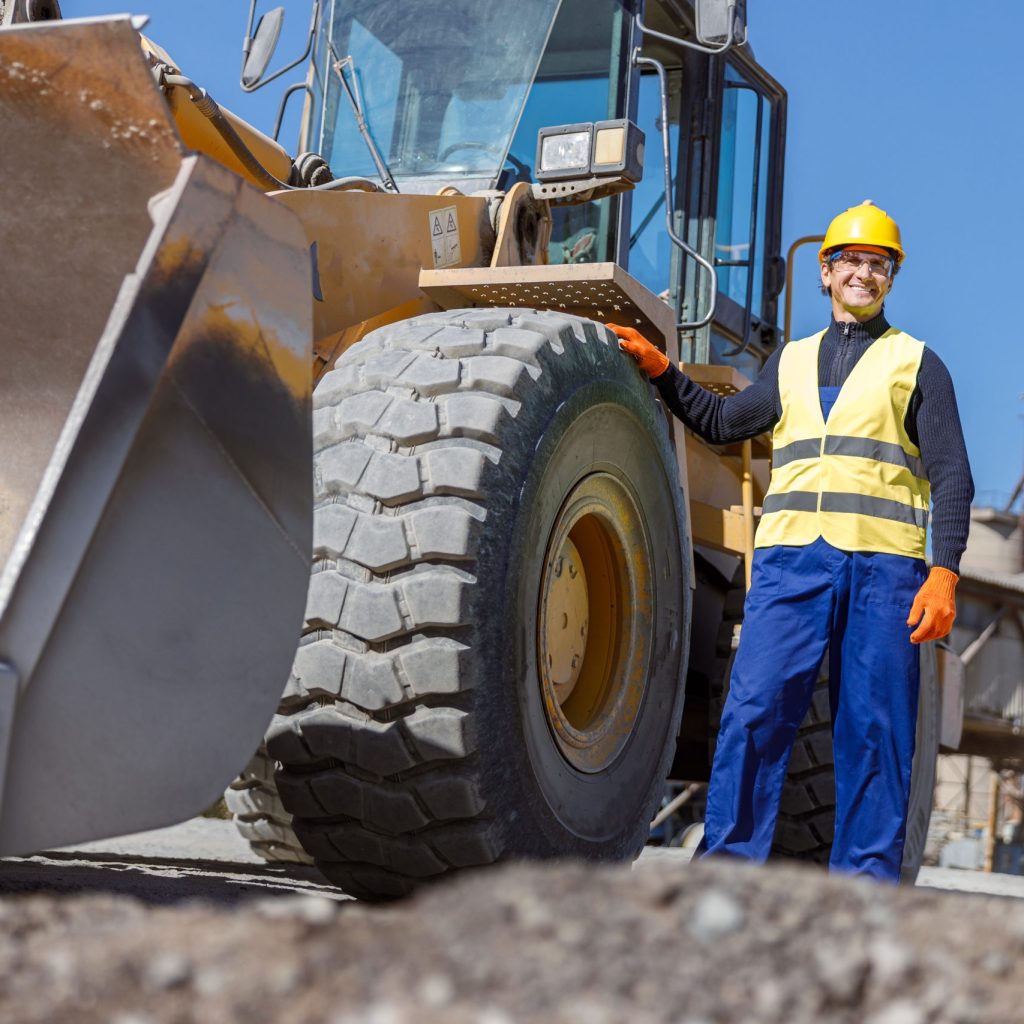 Joyful matured man factory worker placing hand on wheel and smiling while standing near tractor